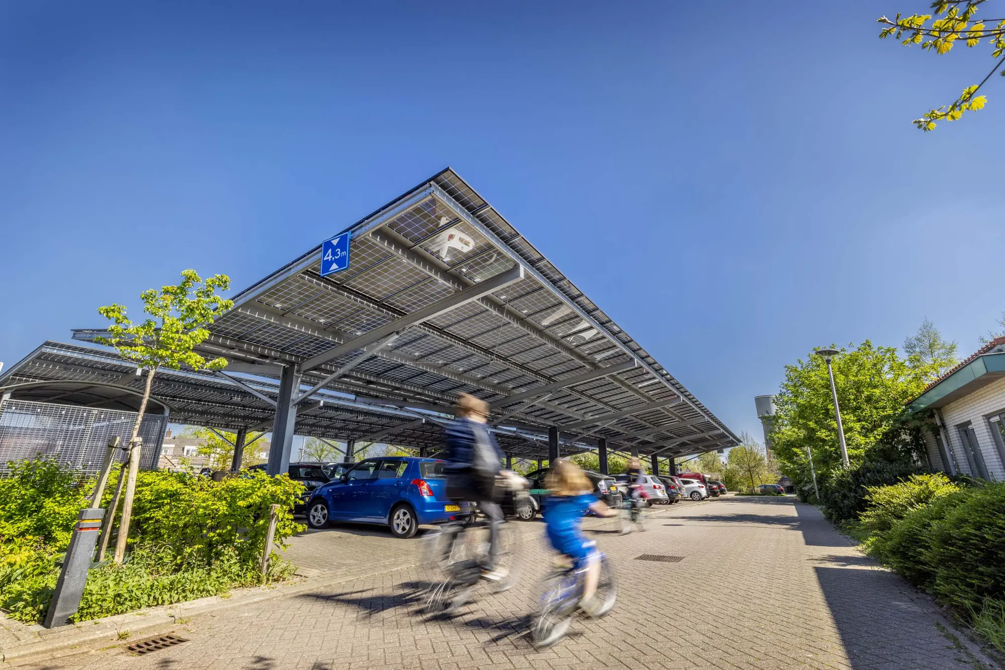Zonnige parkeerplaats overkapt met grote stalen constructies vol zonnepanelen. Op de voorgrond fietsen twee kinderen over de klinkers, terwijl er verschillende auto's onder de overkapping geparkeerd staan.