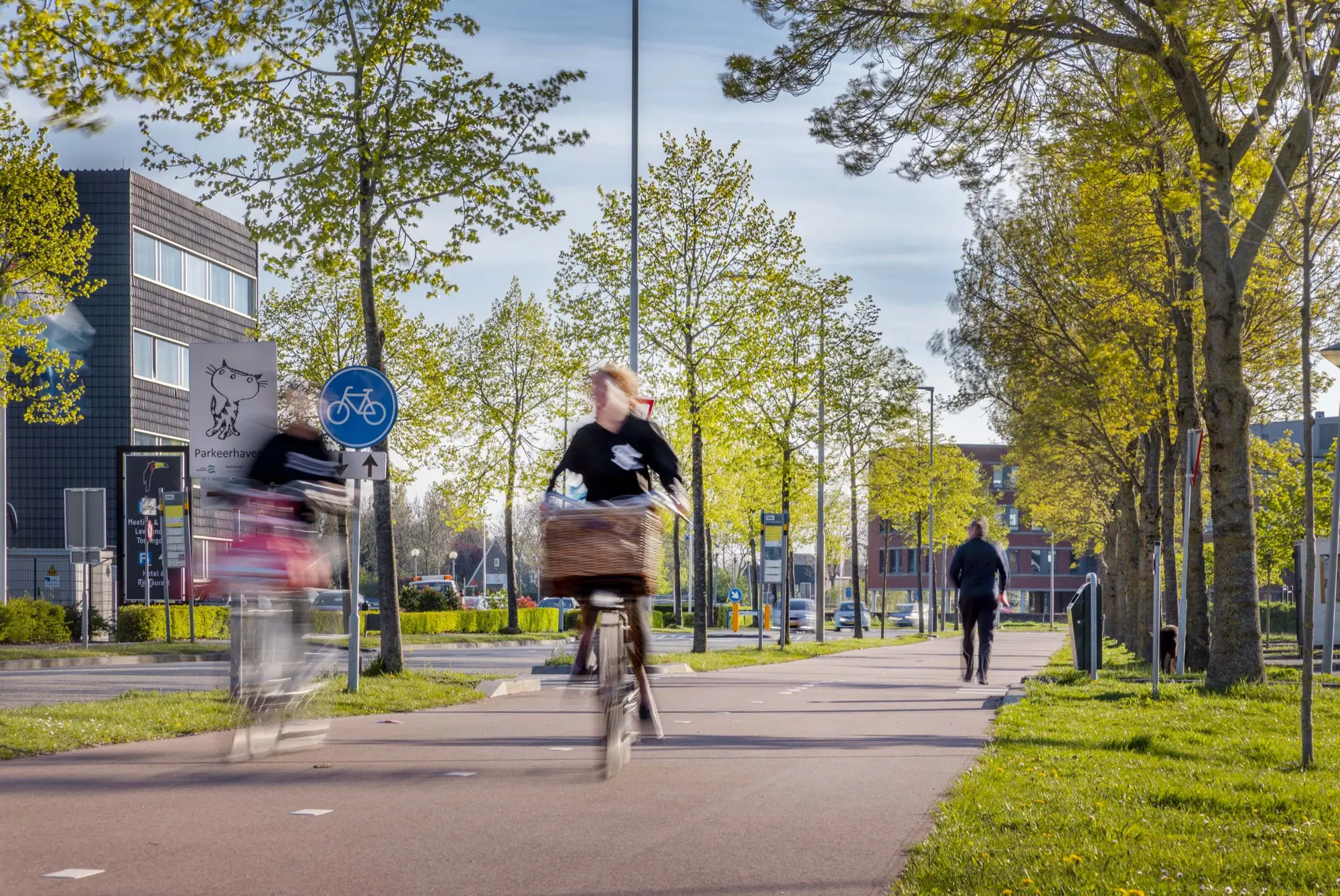 Twee fietsers met bewegingsonscherpte rijden over een breed fietspad met bomen. Links staat een blauw rond bord voor een fietspad en een kantoorgebouw; rechts loopt een voetganger langs het pad.