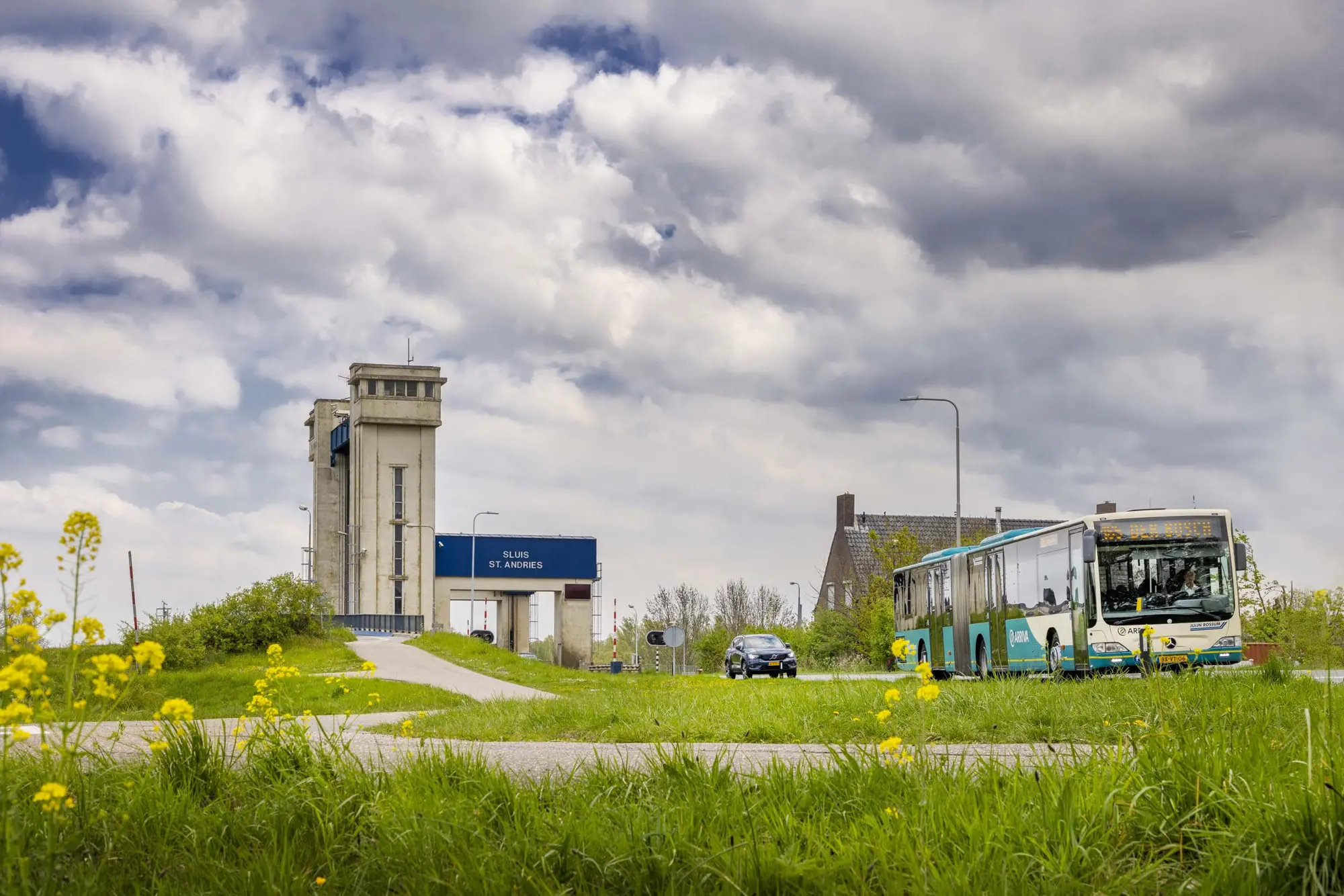 Groene Arriva-lijnbus en een personenauto rijden over een weg langs een groen grasveld met gele bloemen. Op de achtergrond is het bedieningsgebouw en de toren van Sluis St. Andries te zien onder een bewolkte lucht.