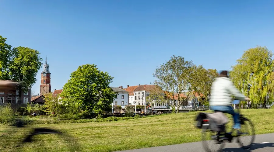 Een fietser met bewegingsonscherpte rijdt op een geasfalteerd pad over een groen talud. Op de achtergrond ligt een historisch stadje aan het water met een opvallende kerktoren onder een helderblauwe lucht.
