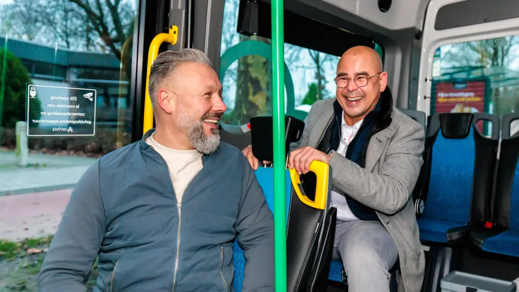 Twee mannen zitten vrolijk lachend en met elkaar pratend op de blauwe passagiersstoelen binnen in een moderne bus.