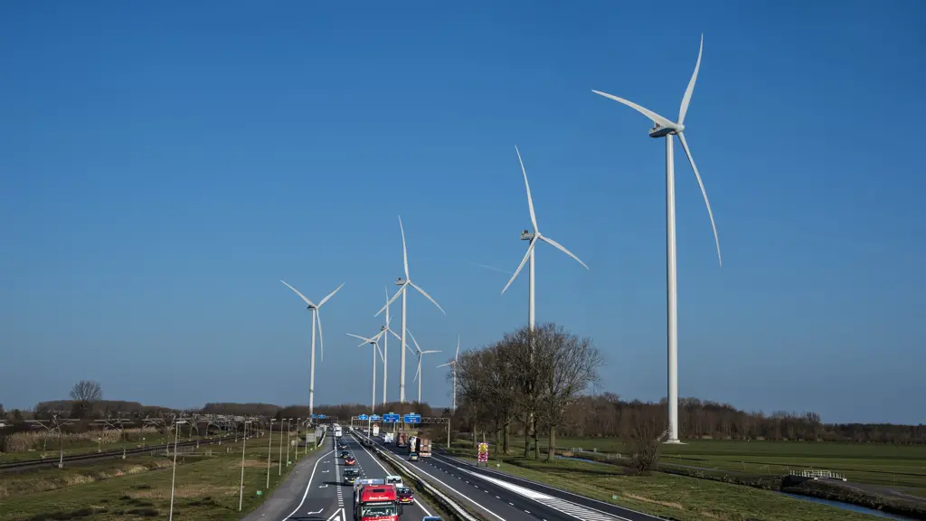 Zicht over een snelweg met rijdend verkeer, waaronder een rode vrachtwagen, waarlangs een lange rij grote witte windturbines staat onder een helderblauwe lucht.