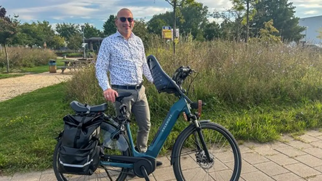 Glimlachende man met zonnebril en een wit overhemd staat op een tegelpad naast een blauwgroene elektrische fiets met dubbele fietstas en een kinderzitje voorop, met weelderig groen op de achtergrond.