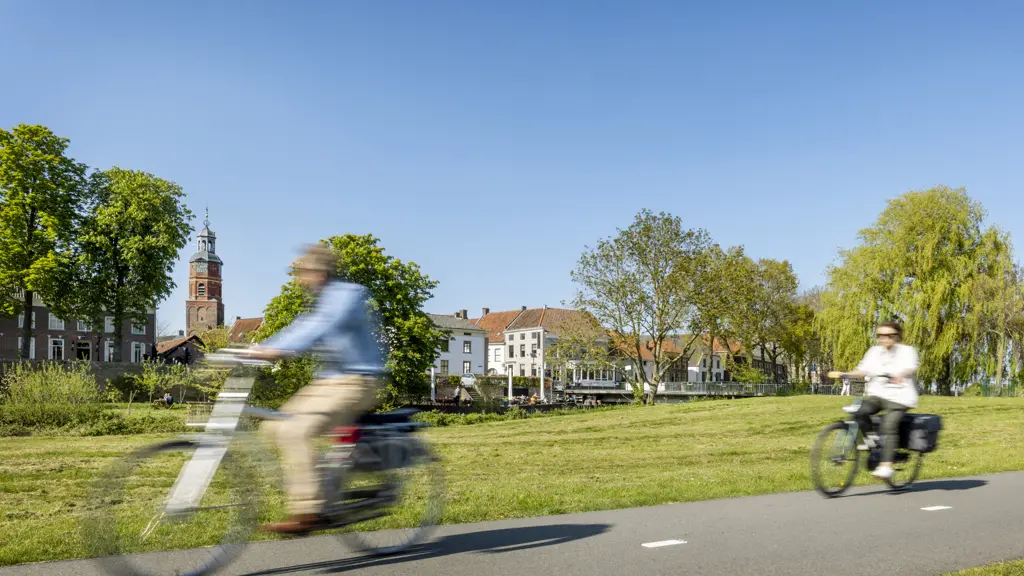 Twee fietsers met bewegingsonscherpte rijden op een geasfalteerd fietspad langs een breed groen grasveld, met op de achtergrond een historisch stadje met een kerktoren en bomen onder een strakblauwe hemel.