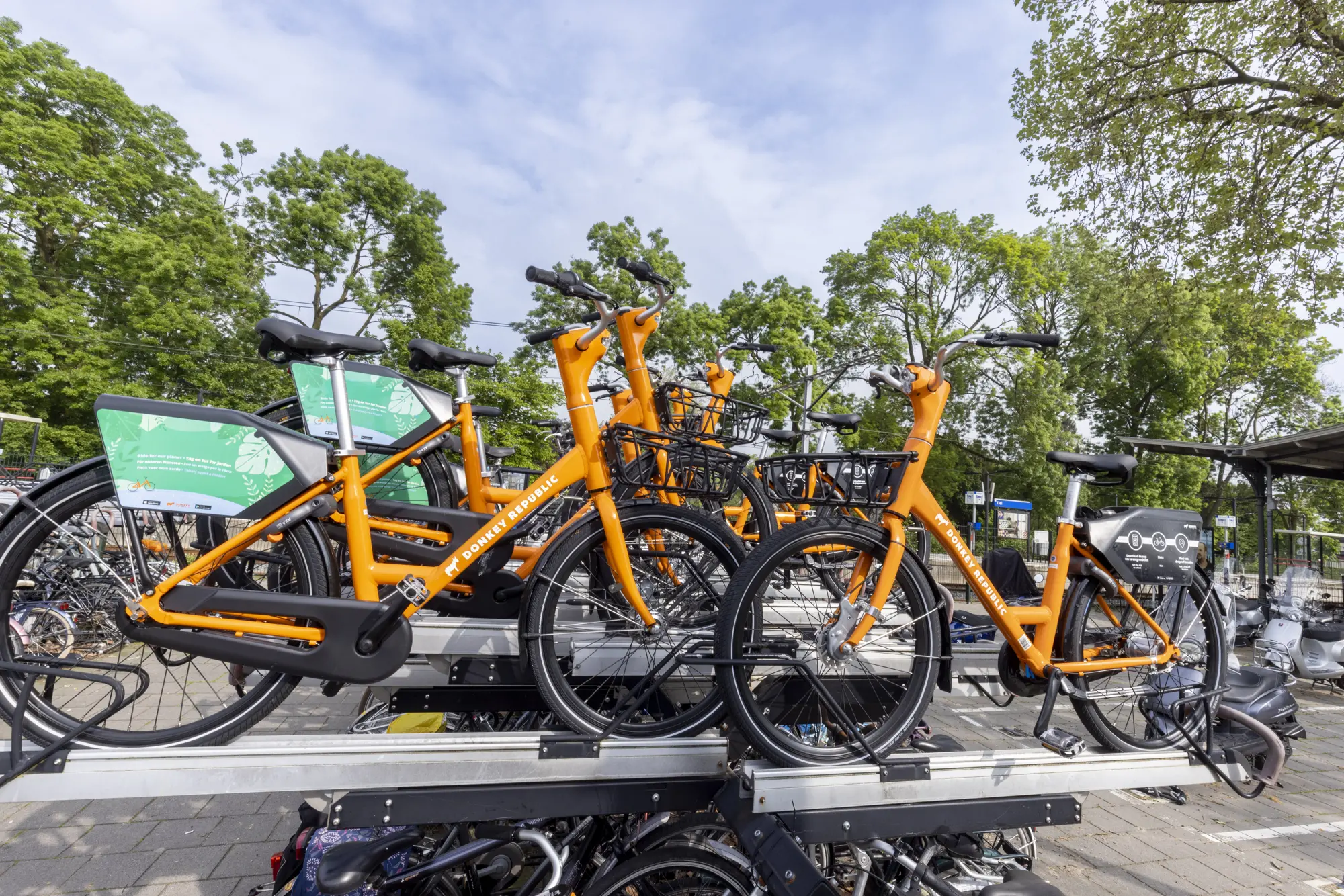 Een stapel van drie oranje Donkey Republic deelfietsen op een aluminium fietsenrek, met op de achtergrond bomen en een treinstation.
