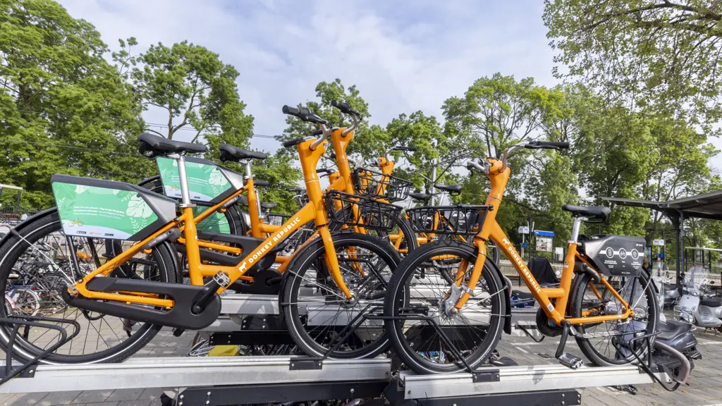 Een stapel van drie oranje Donkey Republic deelfietsen op een aluminium fietsenrek, met op de achtergrond bomen en een treinstation.