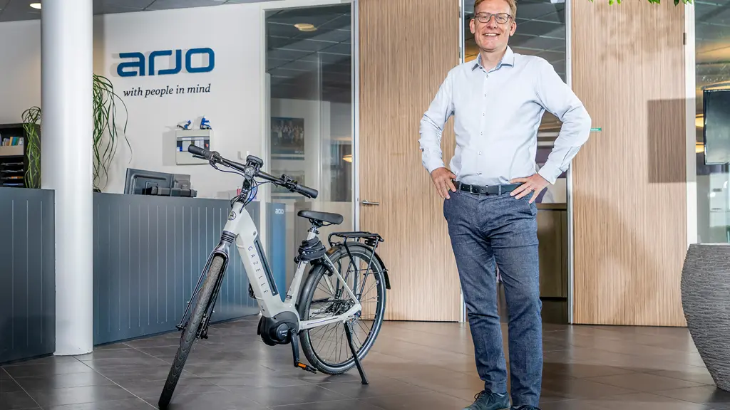Afbeelding van een glimlachende man in een kantooromgeving die poseert met zijn handen op zijn heupen. Hij draagt een lichtblauw overhemd, een donkere broek en blauwe schoenen. Naast hem staat een zilvergrijze Gazelle e-bike op een donkere tegelvloer. In de achtergrond is een receptiebalie te zien met het Arjo-logo en de tekst 'met mensen in gedachten' op de muur.