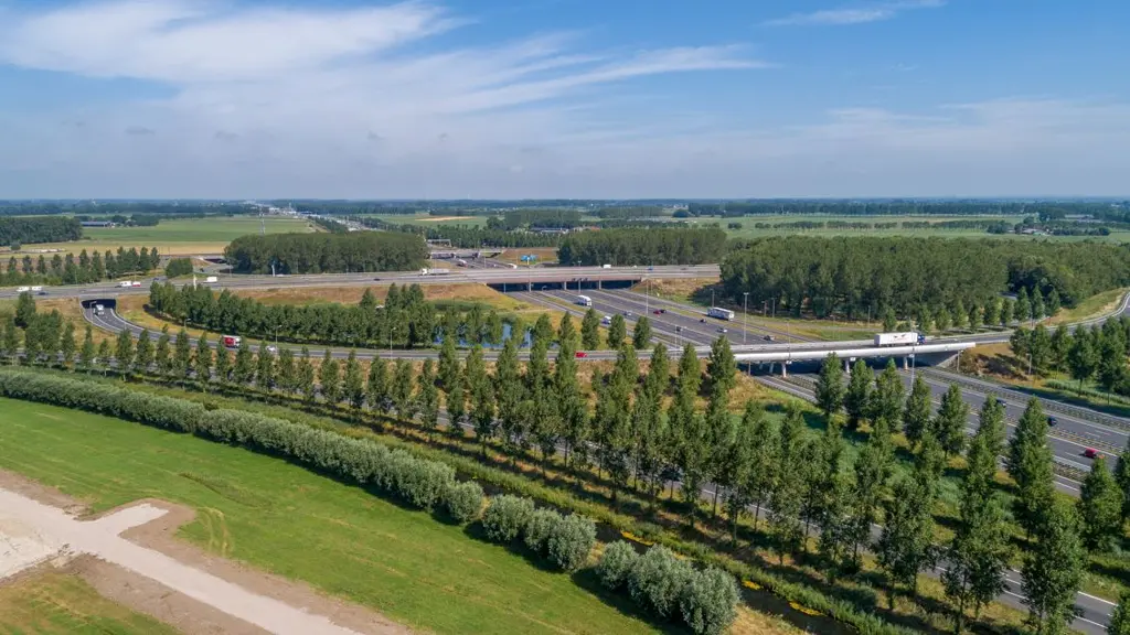 Luchtfoto van een knooppunt van autosnelwegen in een groen landschap. Te zien zijn meerdere wegen, viaducten, groene taluds met rijen bomen en weilanden onder een blauwe hemel met lichte bewolking.