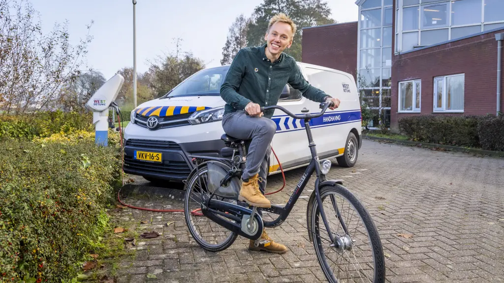 Glimlachende man in een groen overhemd en grijze broek fietst op een zwarte stadsfiets op een klinkerpad. Op de achtergrond staat een witte Toyota Proace van de gemeente West Betuwe met 'Handhaving' op de zijkant, geparkeerd bij een wit oplaadstation. Rechts een modern kantoorgebouw met rode bakstenen en een glazen pui.