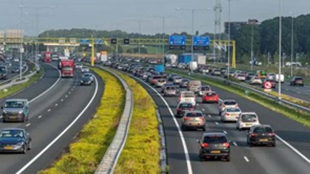 Overzichtsfoto van een drukke autosnelweg met meerdere rijstroken vol auto's en vrachtwagens die in een file staan. In het midden is een strook met groen-gele beplanting. Er staan matrixborden boven de weg.