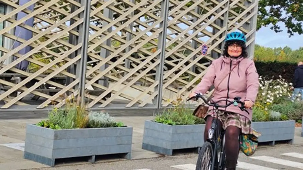 Vrouw in een roze jas en een blauwe helm fietst over een zebrapad op een zwarte fiets met een tas voorop. Op de achtergrond staat een gebouw met een opvallende houten gevelconstructie.
