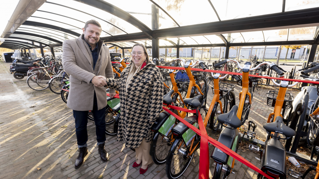 Man in een beige jas en vrouw in een pied-de-poule jas schudden elkaar de hand in een fietsparkeerstalling. Tussen hen in staat een rij oranje deelfietsen van Donkey Republic, met een rood lint dat door de fietsen is geregen. De zon schijnt door het glazen dak van de stalling.