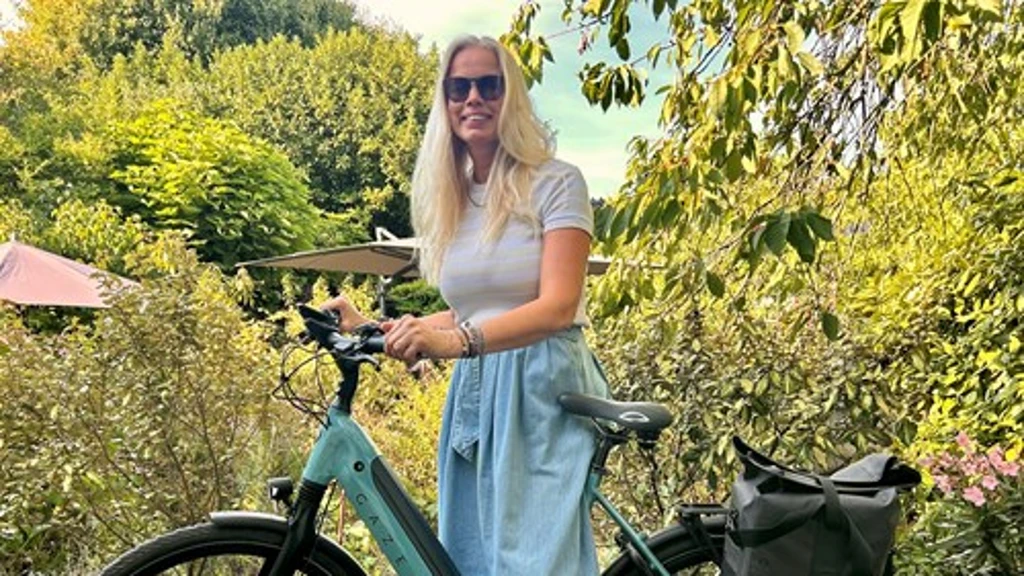 Blonde vrouw met een zonnebril, wit T-shirt en een lichtblauwe culotte staat glimlachend in een weelderige groene tuin naast een mintgroene Gazelle e-bike met een zwarte fietstas achterop.