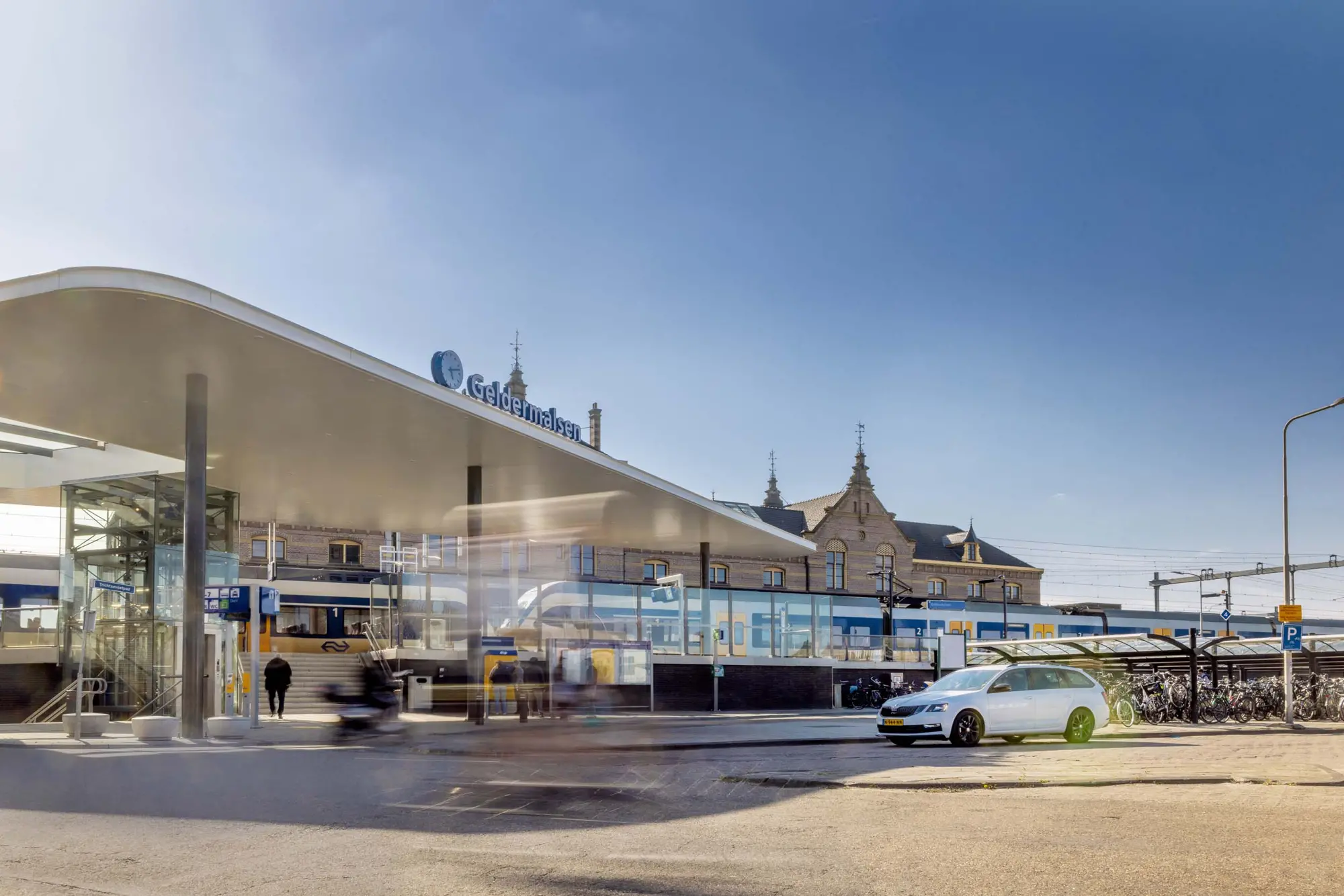 Zijaanzicht van station Geldermalsen met een moderne overkapping en een glazen lift. Op het spoor staat een NS-sprinter en op de voorgrond staat een witte stationwagen geparkeerd op het stationsplein onder een helderblauwe hemel.